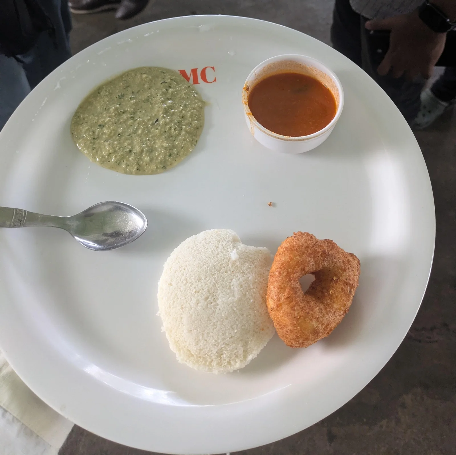 Two idlis and a medu vada on a white plate, with green chutney and a cup of sambar.