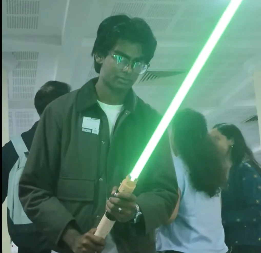 Me holding a lightsaber with a pale wooden hilt and a bright green blade, photographed from below under the venue's ceiling lights.